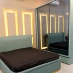 Luxury aqua blue upholstered bed with gold accents, paired with a glass wardrobe featuring automatic lighting for a stylish bedroom setup